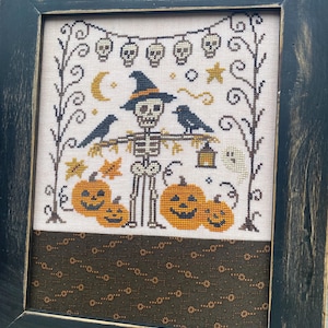以下が含まれることがあります： 黒いアンティーク調のフレームに入ったクロスステッチアート。魔女の帽子をかぶった骸骨、カラス、カボチャ、幽霊、頭蓋骨のガーランドが特徴です。カラーパレットは黒、オレンジ、クリーム色です。