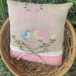 May include: A beige cross-stitched pillow with the word "MOTHER" in pink. The pillow features embroidered birds, flowers, and hearts. A pink polka-dot fabric with lace trim and a twine bow adorns the bottom. The pillow is in a woven basket.