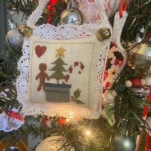 May include: A Christmas tree ornament with a cross-stitch design. The ornament features a small, square pillow with a Christmas tree, a gingerbread person, and a candy cane. The pillow is trimmed with white lace and small gold bells.