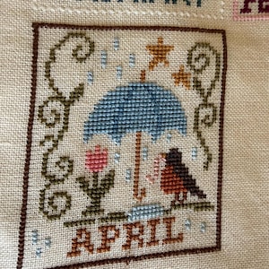 May include: Cross-stitch embroidery featuring the month of April. The design includes a blue umbrella, a red bird, a pink flower, and the word "APRIL" in brown. The background is a light beige fabric with the word "JANUARY" visible.