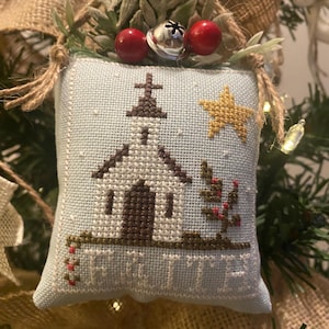 May include: A light blue, cross-stitched Christmas ornament featuring a church, star, and text. The ornament is adorned with red berries, a silver bell, and greenery. The ornament is hanging on a Christmas tree.