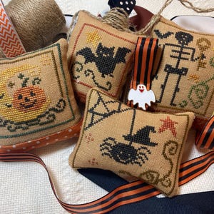 Peut inclure: Plusieurs petits coussins brodés sur le thème d'Halloween. Chaque coussin présente un motif différent, notamment une citrouille, une chauve-souris, une araignée et un squelette. Ruban rayé orange et noir et ficelle sont également présents.