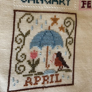 May include: Cross-stitch embroidery featuring the month of April. The design includes a blue umbrella, a bird, a pink flower, and the word "APRIL" in brown. The background is a light beige fabric.