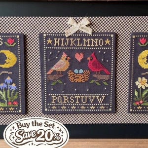 May include: A framed set of three cross-stitch samplers. Each sampler features a dark blue background with embroidered letters, birds, moons, stars, and flowers. The samplers are decorated with bows at the top. The text "Buy the Set Save 20%" is at the bottom.