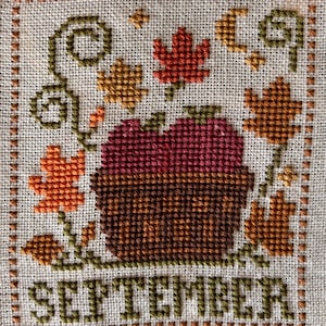 May include: A cross-stitch embroidery on light beige fabric featuring a central brown basket filled with red apples. The design is surrounded by orange and red autumn leaves, green vines, and a crescent moon. The word SEPTEMBER is stitched in green.