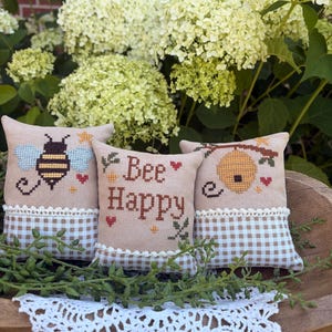 Könnte beinhalten: Drei kleine quadratische Stoffkissen mit Kreuzstichstickerei. Das linke Kissen zeigt eine Biene, das mittlere trägt die Aufschrift Bee Happy und das rechte einen Bienenstock. Jedes Kissen hat eine beige Oberseite, eine braun-weiß karierte Unterseite.