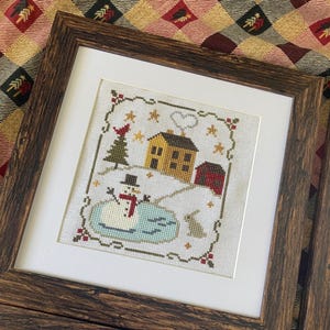 May include: Framed cross-stitch artwork featuring a winter scene. The design includes a yellow house, red barn, snowman, and a small pond. The artwork is framed in a dark brown, textured wooden frame.
