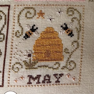Puede incluir: Bordado de punto de cruz con un diseño de mayo, que incluye una colmena, abejas y detalles florales. La palabra "MAY" está bordada en marrón. El diseño está enmarcado con puntadas decorativas sobre un fondo de tela neutra.