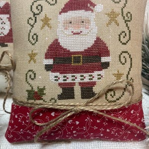 Peut inclure: Un petit coussin de Noël carré avec un motif de Père Noël brodé au point de croix. Le coussin a un fond beige avec une section inférieure rouge et est noué avec de la ficelle. Le motif du Père Noël comprend un costume rouge, une barbe blanche et un visage souriant.