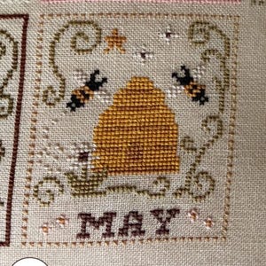 May include: Cross-stitch embroidery featuring a May design with a beehive, bees, and floral accents. The word "MAY" is stitched in brown. The design is framed with decorative stitching and set against a neutral fabric background.
