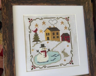 May include: Cross-stitch artwork in a wooden frame, depicting a winter scene with a snowman, house, and barn. The image shows a snowman, a yellow house, a red barn, and a green Christmas tree, all framed with a white mat.