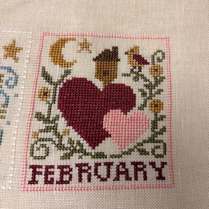 May include: Cross-stitch embroidery featuring a February design. The design includes a large burgundy heart, a smaller pink heart, a house, a bird, and the word "FEBRUARY" in burgundy. The design is framed in pink and set against a beige fabric background.