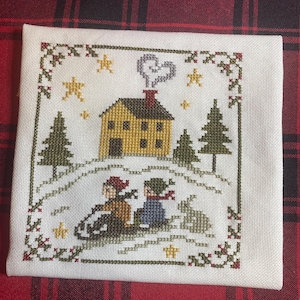 May include: Cross-stitch embroidery featuring a winter scene with a yellow house, two figures sledding, and a rabbit. The design includes green trees, stars, and a decorative border with red and green accents. The background is white.