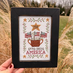 May include: Framed cross-stitch art with a black wooden frame. The artwork features a patriotic design with American flags, stars, and the text "1776-2026 AMERICA". The central element is a woven basket holding three American flags.