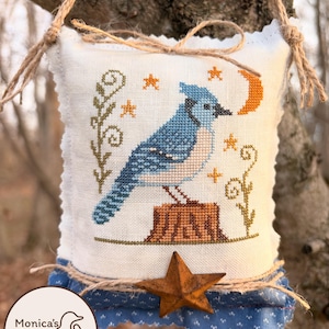 May include: A handmade, square, off-white fabric ornament with a blue jay cross-stitch design. The bird is perched on a tree stump, with a crescent moon and stars. A rusty star and blue fabric trim adorn the bottom. The ornament is suspended by twine.