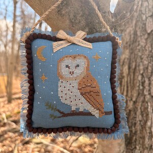 May include: A square, blue cross-stitch pillow featuring a brown and white owl perched on a branch. The pillow has a brown pom-pom trim, a frayed edge, and a small bow at the top. A jute rope is attached for hanging.