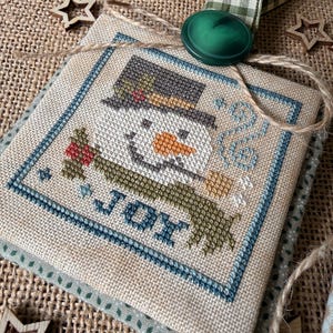 May include: A square fabric ornament featuring a cross-stitched snowman wearing a dark hat and a green scarf, with the word JOY stitched in blue. The ornament is topped with a dark green button and a green and white checkered ribbon loop.
