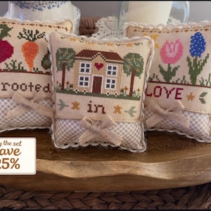 May include: Three decorative, small, square pillows with cross-stitch designs. One pillow features a radish and carrot with the word "rooted". Another has a house with the words "in". The third pillow has flowers and the word "love". Each pillow has a bow.