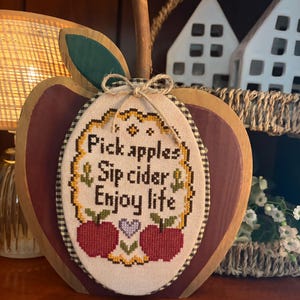 May include: A wooden apple-shaped cutout with a dark red finish and a green leaf, featuring a central oval cross-stitch embroidery. The embroidery displays the text "Pick apples, Sip cider, Enjoy life" with two red apples and a small purple heart.
