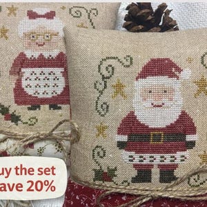 May include: Two Christmas-themed decorative pillows with cross-stitched designs. One features Santa Claus, the other Mrs. Claus. Both have a beige background, red accents, and are tied with twine. The pillows are decorated with stars and greenery.