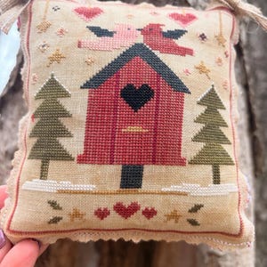 May include: A cross-stitched, square ornament featuring a red birdhouse with a black heart-shaped opening, flanked by green trees. Pink birds and hearts decorate the beige background. The ornament is trimmed with a beige lace and has a jute string for hanging. The text "Monica's Stitching" is in the bottom right corner.
