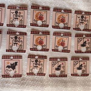 Könnte beinhalten: Eine Sammlung von sechzehn quadratischen Garnspulen aus Pappe mit Halloween-Motiven wie Fledermäusen, Skeletten, Kürbissen und Spinnen. Jede Spule hat einen rot-schwarzen Karorand, ein Loch an der Oberseite und eine Aussparung an der Unterseite.
