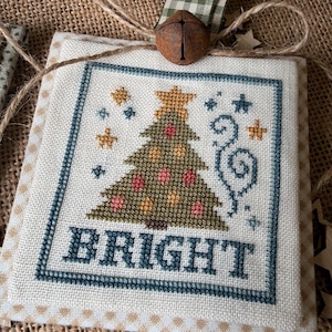 May include: A square cross-stitch ornament on cream fabric featuring a green Christmas tree with red and yellow ornaments, a yellow star topper, and the word BRIGHT in dark blue. It includes a green gingham ribbon, a rustic metal bell, and twine.