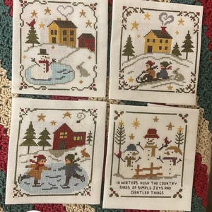 May include: Four cross-stitch winter scene panels. Each panel features a different winter scene with houses, snowmen, and people ice skating. The panels are framed with a decorative border and are on a cream-colored fabric.