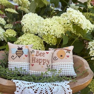 May include: Three small square fabric pillows featuring cross-stitch embroidery. The pillows have a beige top section with bee-themed designs and text reading Bee Happy, and a bottom section with a white and beige gingham pattern and lace trim.