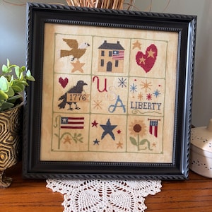 May include: Framed cross-stitch sampler with a black frame. The sampler features patriotic symbols like an eagle, house, heart, stars, and flags. The word "LIBERTY" is stitched in blue, and the year "1776" is included. The background is a neutral beige.