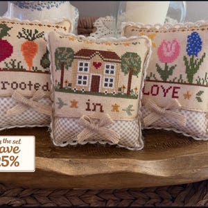 Puede incluir: Tres pequeñas almohadas cuadradas decorativas con diseños de punto de cruz. Una almohada presenta un rábano y una zanahoria con la palabra "rooted". Otra tiene una casa con las palabras "in". La tercera almohada tiene flores y la palabra "love". Cada almohada tiene un lazo.