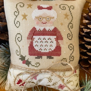 May include: A decorative pillow featuring a cross-stitched design of a woman in a red dress and white apron. The pillow has a beige background with a floral pattern at the bottom and is tied with twine. The design includes stars and holly.