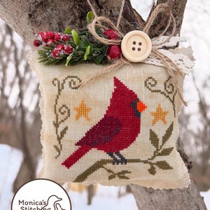 May include: A decorative, square, cross-stitched pillow featuring a red cardinal bird perched on a branch, with yellow stars and green leaves. The pillow is adorned with a button, faux berries, and greenery. The pillow is from Monica's Stitching Studio.