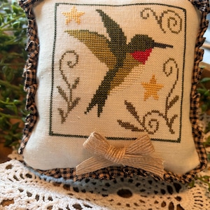 Puede incluir: Pequeño cojín decorativo cuadrado con un diseño de colibrí bordado a punto de cruz. El ave es verde y roja, con estrellas amarillas y detalles florales. Tiene un borde de cuadros vichy con volantes y un lazo de yute.