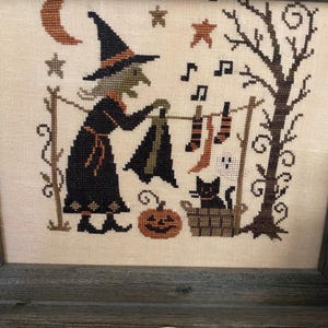 May include: Cross-stitch artwork in a wooden frame depicts a witch hanging laundry on a clothesline. The witch is wearing a black dress and hat. A black cat sits in a basket, and a jack-o'-lantern is on the ground. Stars, a moon, and musical notes are also included.