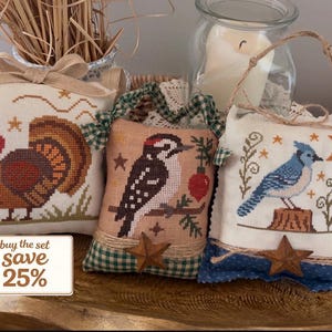 May include: Three decorative, cross-stitched pillows with bird and nature designs. One features a turkey, another a woodpecker, and the third a blue jay. Each pillow has a star accent and a rustic, homespun aesthetic. The text "buy the set save 25%" is visible.