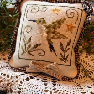 May include: A square, off-white decorative pillow with a cross-stitched hummingbird design in shades of green and brown, accented with yellow stars and foliage. The pillow has a black and tan gingham border and a twine bow.