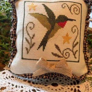 May include: A small, square decorative pillow with a cross-stitched hummingbird design. The bird is green and red, with yellow stars and floral accents. The pillow has a ruffled gingham border and a jute bow.