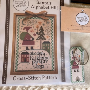 May include: A cross-stitch pattern titled Santa's Alphabet Hill by Monica's Stitching Studio, featuring a Santa Claus figure, a pine tree, houses, and the alphabet. The set includes a printed pattern sheet and a small ring of paper tags.