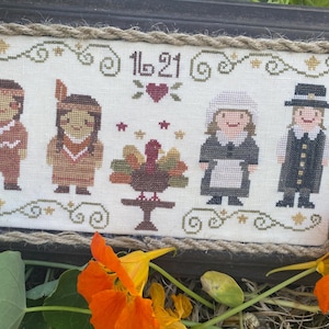 May include: Cross-stitch artwork in a dark wooden frame with a rope border. The artwork depicts four figures: two Native Americans, a woman in a white bonnet and apron, and a man in a black suit. A turkey sits on a table. The text "16 21" is above the figures.
