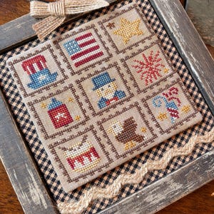 May include: Framed cross-stitch sampler with patriotic motifs. The sampler features nine squares with images of the American flag, a star, fireworks, a top hat, a firecracker, an eagle, and a drum. The frame is distressed gray wood, and a jute bow adorns the top.