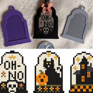 May include: Three tombstone-shaped plastic frames in purple, black, and gray, with a grid pattern. The black frame features a cross-stitch design with the text "OH NO" and a skull. Below are three cross-stitch patterns: a skull with "OH NO" text, a black cat with a pumpkin, and a haunted house.
