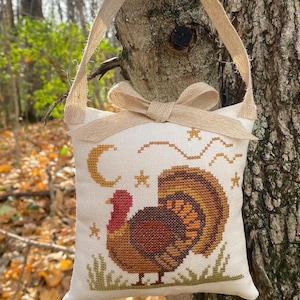 May include: A cross-stitched Thanksgiving-themed ornament featuring a turkey, crescent moon, and stars. The ornament has a beige background with a brown turkey, and a beige ribbon for hanging. The bow is tied at the top.