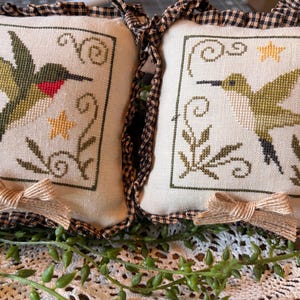 May include: Two square decorative pillows with cross-stitched hummingbird designs. The hummingbirds are in shades of green, white, and red, with yellow stars and floral accents. Each pillow has a black and white checkered ruffle and a burlap bow.