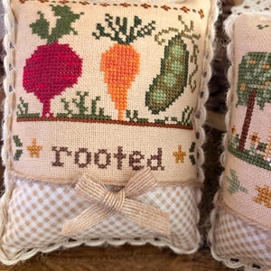 May include: A small, square, cross-stitched pillow featuring a red beet, orange carrot, and green pea pod. The word "rooted" is stitched below the vegetables. The pillow has a beige gingham base and a jute bow.