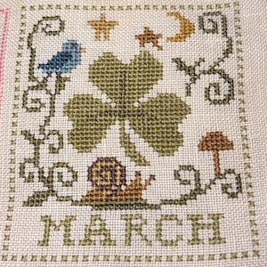 May include: Cross-stitch design featuring a four-leaf clover, bluebird, snail, stars, and moon. The word "MARCH" is stitched at the bottom. The design is framed with a green border and stitched on a cream-colored fabric.