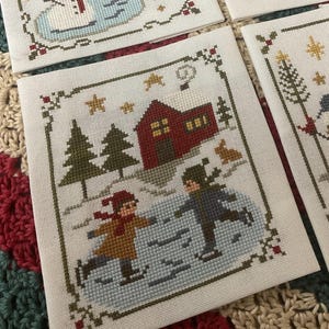 May include: Cross-stitch winter scene featuring two children ice skating near a red house and evergreen trees. The design includes a border with stars and floral accents. The color palette includes red, brown, green, and blue.