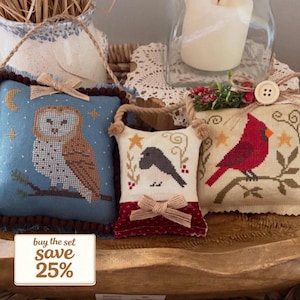 May include: Set of three decorative, cross-stitched pillows. One features an owl on a blue background, another has two birds, and the third displays a red cardinal. Each pillow has a bow and a hanging loop.