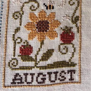 May include: A cross-stitch embroidery on light fabric featuring a central yellow sunflower with a dark brown centre, two red berries, a small black bee, and a crescent moon. The word AUGUST is stitched in dark brown letters at the bottom.