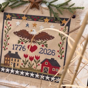 May include: Cross-stitch artwork on burlap fabric, featuring an eagle, stars, and the years 1776 and 2026. The design includes a red heart, houses, and floral elements, framed with a rustic wooden border and decorative accents.